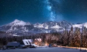 Zimowe Tatry nocą, Droga Mleczna nad ośnieżonymi szczytami i lasem. W dole przytulne górskie domki, idealne miejsce, gdzie w góry zima zaprasza.