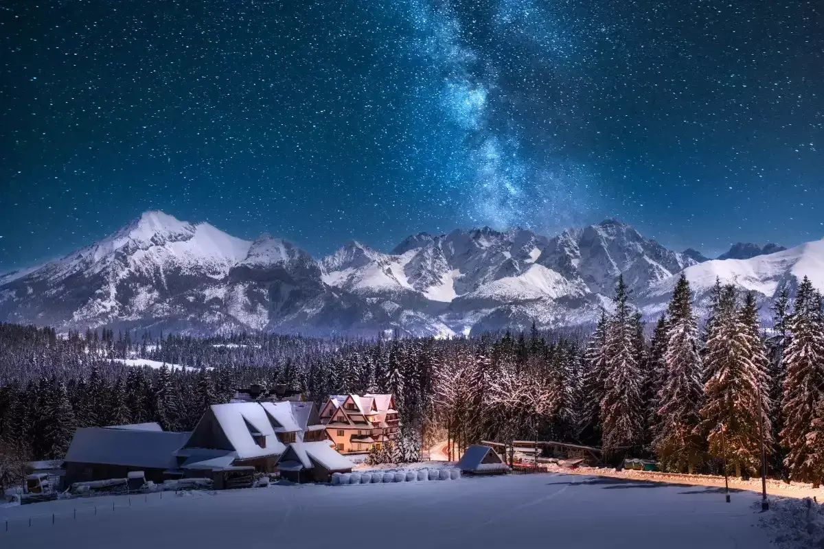 Zimowe Tatry nocą, Droga Mleczna nad ośnieżonymi szczytami i lasem. W dole przytulne górskie domki, idealne miejsce, gdzie w góry zima zaprasza.
