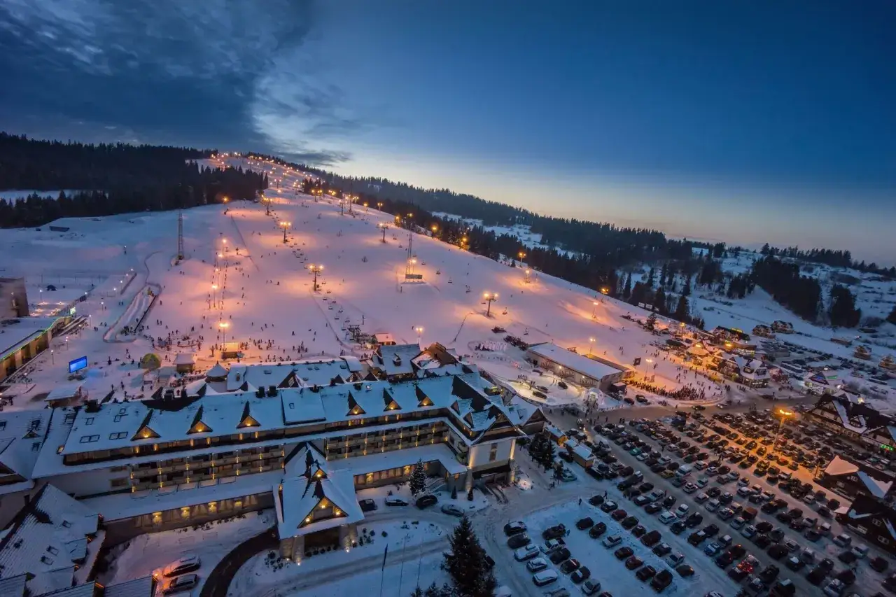 Oświetlony stok narciarski w Białce Tatrzańskiej nocą, z hotelem i parkingiem. Mnóstwo atrakcji dla narciarzy i snowboardzistów.
