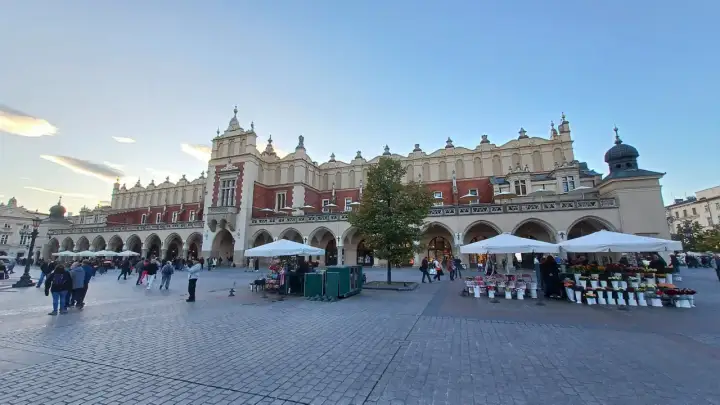 Sukiennice, jeden z najpiękniejszych zabytków w Krakowie, tętni życiem. Ludzie spacerują po Rynku Głównym, podziwiając architekturę i kupując kwiaty.